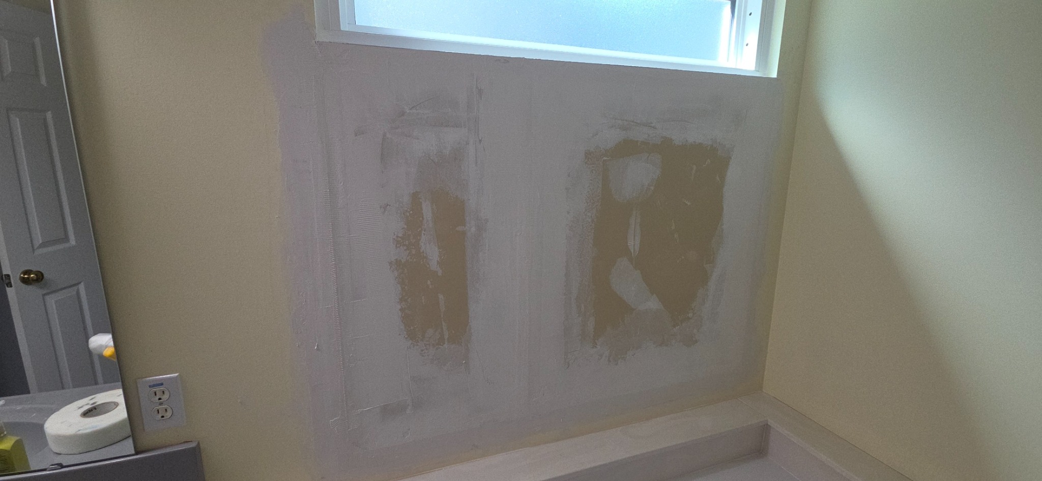 Bathroom wall before repair with visible drywall damage and patches beneath the window